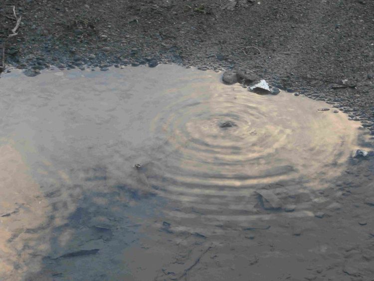 Water from an underground leak welling up through a puddle on gravel, rippling out from one point